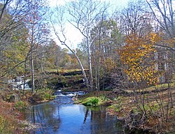 A creek with some waterfalls over an old dam in the distance on a clear autumn day. There is a house on the right obscured by the trees