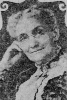 An older white woman with grey hair in an updo, wearing glasses and a lace-trimmed high-collared dress, her cheek resting on one hand