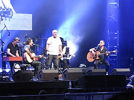 A stage shot with four men clearly visible. The man at left is leaning at a rectangular box, he has a microphone nearby. The second man is shown in right profile, he is playing a guitar and singing into a microphone while seated. The third man is standing in the middle of the stage with arms at his sides, he carries a microphone in his right hand. The fourth man is also seated while playing a guitar and at a microphone. A fifth man is obscured seated behind and to the right of the middle one. The men are surrounded by stage equipment including speakers, lights and additional microphones. A drum kit is partly visible but the drummer is obscured.