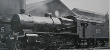 Side-and-front view of a modified version of the N class on shed. The distinguishing feature from normal N class locomotives is the tall cylindrical chimney on the smokebox. A member of the crew is standing next to the tender.