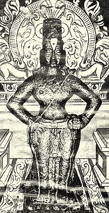 Черно-белое изображение идола вооруженного акимбо мужчины с обнаженной грудью, в коническом головном уборе, дхоти и украшениях. Идол помещен на кирпич и окружен украшенным нимбом.