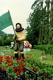 Photographie en couleur d'une statue de Tintin mise en scène dans un parc. Il porte un drapeau vert siglé F.E.R.S. et des champignons rouge et blanc sont installés derrière lui.