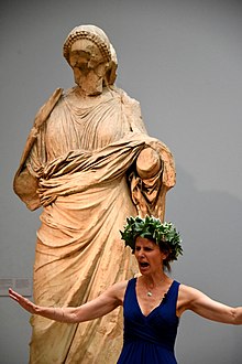 An actress performing a play. She wears a laurel wreath and stands in front of a statue of a woman from the Mausoleum at Halicarnassus (room 21, The British Museum, London) An actress performing a play. She wears a laurel wreath and stands in front of a statue of a woman from the Mausoleum at Halicarnassus. Room 21, The British Museum, London.jpg
