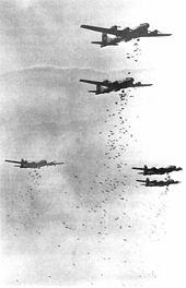 Photographie en noir et blanc d'une formation de cinq quadrimoteurs. Chacun d'eux largue plusieurs dizaines de bombes.