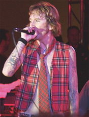 A man wearing an unbuttoned vest and a red tie holds a microphone to his face.