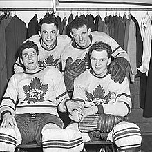 Ice hockey players in a locker room. Two are sitting on a locker room bench, with another two players standing behind them.