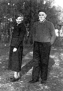 A white woman and a white man, standing outdoors; she is wearing a dark suit, and he is wearing a beret, sweater, and trousers; he has a white beard and mustache, and dark eyebrows