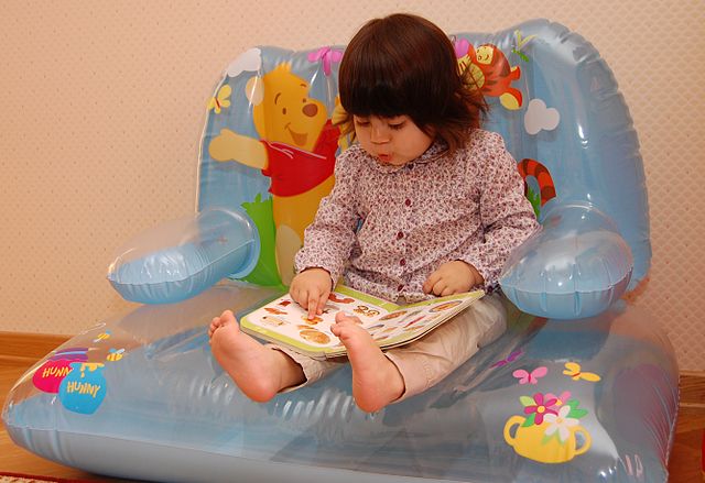 A girl is sitting and looking down at a picture book that rests on her legs, while she rests her finger on an image.