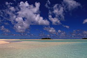 Very shallow water over a white seafloor with a green island in the background beneath blue sky and scattered clouds