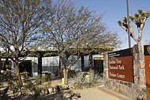 На этом изображении показан вход в Центр для посетителей Национального парка Джошуа-Три в Джошуа-Три, Калифорния. На нем изображен большой коричневый знак с названием центра для посетителей, а также зона отдыха на открытом воздухе с пустынной растительностью, такой как агава и одно растение Джошуа.