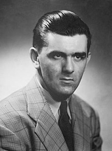 Upper body of a young man in a suit and tie with slicked back hair and a serious look on his face.