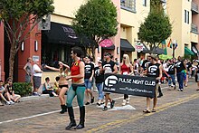 Группа людей в рубашках с логотипом Pulse марширует по кирпичной улице с транспарантом с логотипом клуба. Человек, одетый как Робин из Бэтмена, возглавляет процессию.