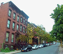 A row of three-story rowhouses in varied colors along a street with small trees and cars parked in front of them, looking uphill