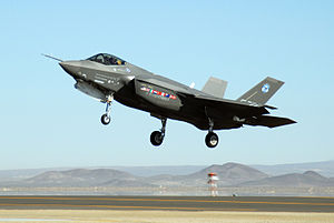 Gray jet aircraft taking off on a clear blue sky, with the landing gear still protruding from its underside. Mountains make-up the background.