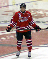 A hockey player, wearing a red Oshawa Generals jersey, stands in full gear on ice with stick held across his waist.