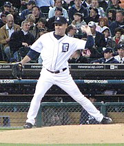 Man in a white baseball uniform stretches to throw a baseball with his left hand