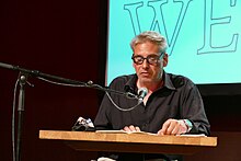 Oberkörper eines sitzenden Mannes mit kurzen grauen Haaren und dickrandiger Brille, der in ein Standmikrofon spricht. Vor ihm ein Holztisch mit einem aufgeschlagenen Buch in das er blickt.