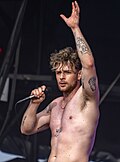 A topless man performs on stage with a microphone and one arm in the air
