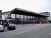 A glass and steel building with a canopy, a row of shops at ground level. There is a bus waiting at a bus stop in front of the building, and cars waiting with people. A sign above an entrance readings "Barking" with symbols for National Rail and London Underground.