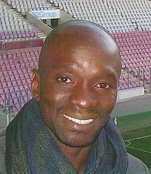 Épaules et visage d'un homme de face. Pose dans une tribune de stade, tribune opposée en arrière-plan.