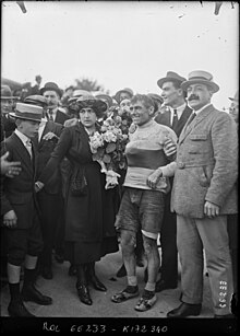 Photographie en noir et blanc d'un cycliste couvert de boue, entouré par la foule, un homme le tenant par le bras.