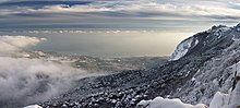 Blick vom Berg ins Tal mit klaren Wolken, die vom Horizont bis zum Berg reichen. Von der Mitte bis zur linken Seite des Fotos erstreckt sich eine Bucht, wo sich ein kleiner Ort mit weißen Gebäuden befindet. Der größte Teil der rechten Fotoecke zeigt den teils schneebedeckten, teils waldbedeckten Berg.