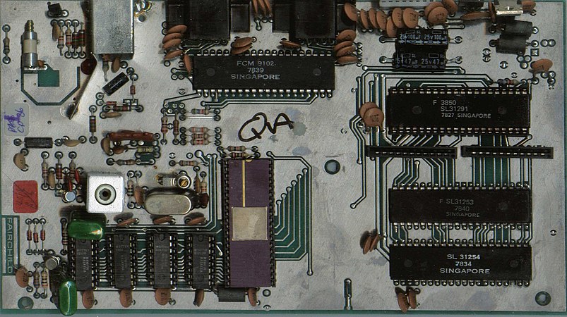 PCB for the derivative Grandstand Video Entertainment Computer. Chips on this board come from Singapore, and a QA mark can be seen. Strangely, most metal on the circuit board has not been etched off, with only areas around the traces having been removed.