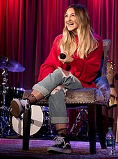 Julia Michaels sitting in a red sweater and jeans on a stage