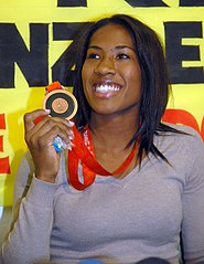 A Brazilian women in casual clothing holding a bronze medal up in her right hand