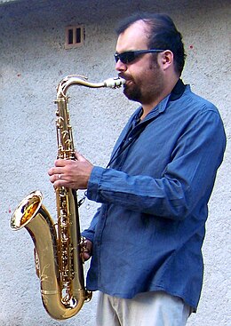 un homme barbu avec lunettes de soleil et chemise bleue en train de jouer du saxophone