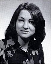 Формальная поза молодой женщины лет двадцати с прямыми темными волосами, разделенными пробором по центру, одетая в темный топ с цветочным принтом.