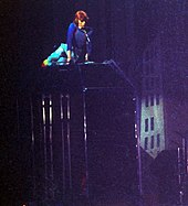 Photo d'un homme aux cheveux roux allongé sur une passerelle dans un décor plongé dans l'obscurité où l'on distingue la silhouette d'un immeuble