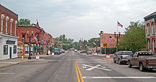 Широкая улица с двойной желтой линией и зданиями по обе стороны, больше слева, чем справа. Американские флаги на фонарных столбах, а знаки как на дороге, так и рядом с ней указывают на железнодорожный переезд вдали.