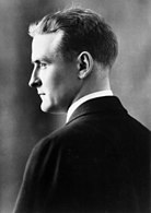 Photograph of F. Scott Fitzgerald in profile circa 1927. His back is towards the camera, and his face is in left profile. He is wearing a dark suit and white shirt. His face has a serious expression as if staring intensely at someone off-camera.