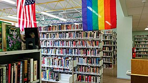 Partial view of the library stacks, with six rows of stacks of bookshelves. Part of a rainbow flag and United States flag are visible hanging from the ceiling.