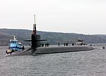 Surfaced submarine in bay with mountains in the background. Flanked to its starboard side by a tug, the boat has several people standing atop of it.