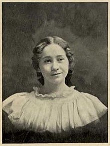 A young white woman wearing a voluminous white blouse; her hair is center-parted and in a low chignon; she is slightly smiling.