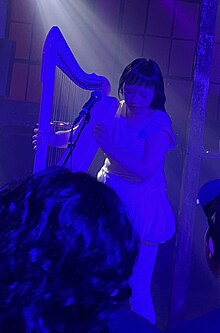 Gao looking down while playing a harp at an indoor live music setting; she is bathed with indigo light