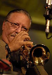 A man wearing glasses and a jacket, playing a trumpet behind a microphone.