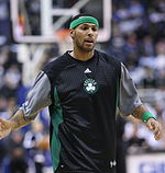 A light-skinned black man is holding out his hand during a basketball pre-game to slap hands with another player. He is wearing a warm-up basketball suit.