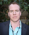 Bret Easton Ellis is pictured standing on front of a stone wall and greenery. He is wearing a blazer over a blue collared shirt and has a lanyard on his neck.