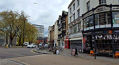 A wide street with old buildings on one side and trees on the other. Tall modern office buildings appear in the distance.