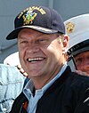 Grammer stands smiling in a white collared shirt, black jacket, and blue baseball cap that reads 'USS Kearsarge' and features the ship's insignia