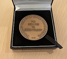 A photograph showing a round bronze medal (the Lord Cohen Medal) in a black gift box. The medal has text around the outside which reads "LORD COHEN OF BIRKENHEAD MEDAL", and in the centre reads "FOR SERVICES TO GERENTOLOGY"