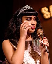 A young, tan-skinned woman with shoulder-length black hair wearing a tiara and silver shirt holds a microphone.