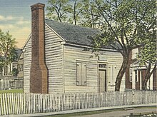 Colorized photo of a log-cabin-style building with a brick chimney and a small tree growing near the door