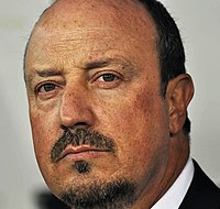 Rafael Benitez is seen with a beard while wearing a coat over a buttoned-up shirt.