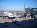 A jet passenger aircraft on the left of the image parked at a glass jetbridge that runs on the right of the image. The plane is surrounded by three aircraft handling cars.