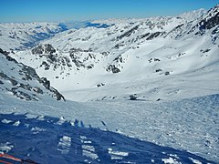 Photographie d'un glacier dans un vallon, avec des sommets enneigés au fond de la photo, le paysage étant enneigé : au centre, la neige est partout hormis sur de hauts rochers noirs qui dépassent ça et là avec un peu d'ombre à gauche de la photo où la montagne du Peyron s'assombrit.