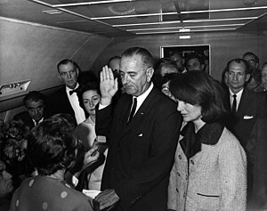 Lyndon B. Johnson giơ tay phía trên cuốn Kinh Thánh dang ra, nhậm chức Tổng thống trong lúc chiếc Không lực Một chuẩn bị rời khỏi sân bay Dallas Love. Jacqueline Kennedy đứng nhìn trong bộ trang phục dính máu (không thấy được).